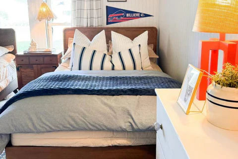 
      A bedroom with two beds, the focus being on one bed with striped pillows and a blue duvet and blanket at the end, as well as a dresser with a plant and orange lamp.
    