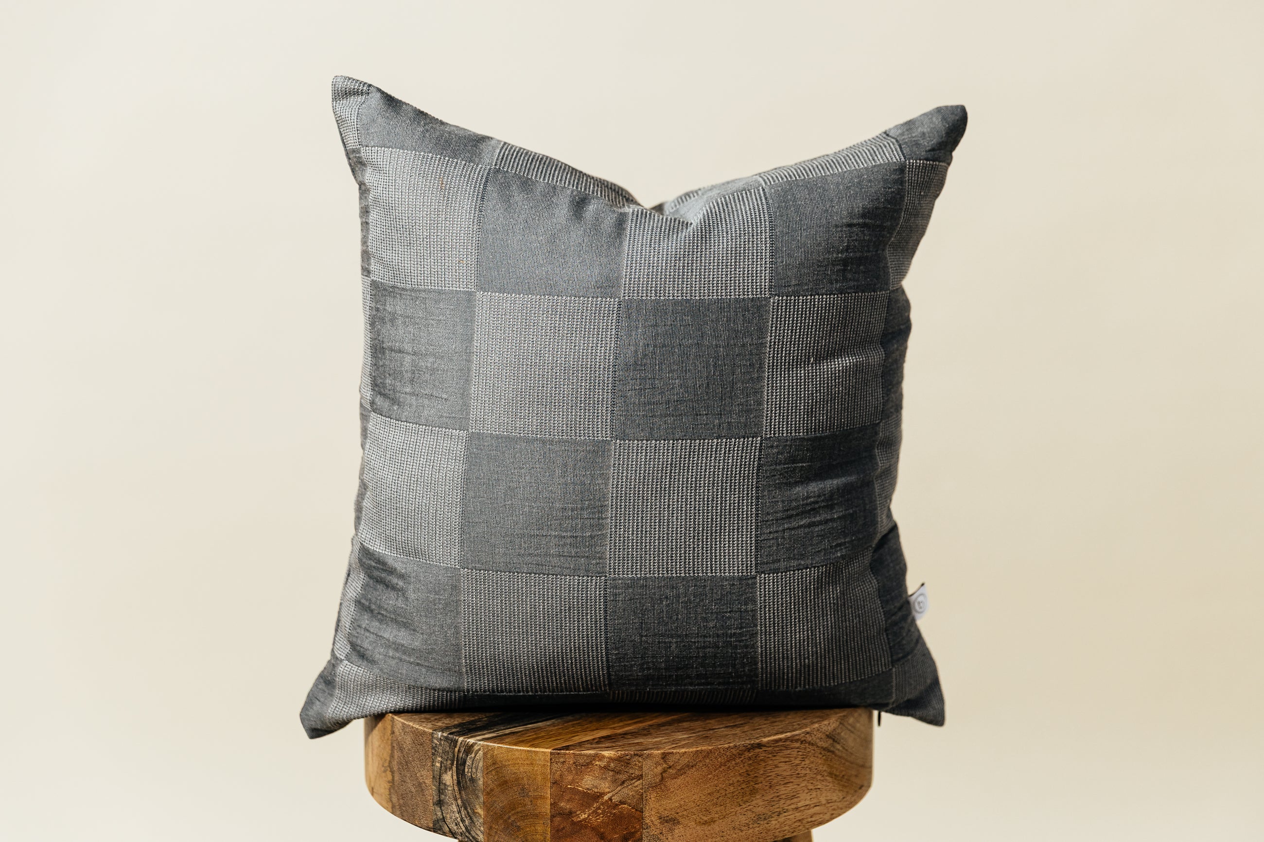 A two toned gray checkerboard pillow sitting on a wooden stool with a cream backdrop.