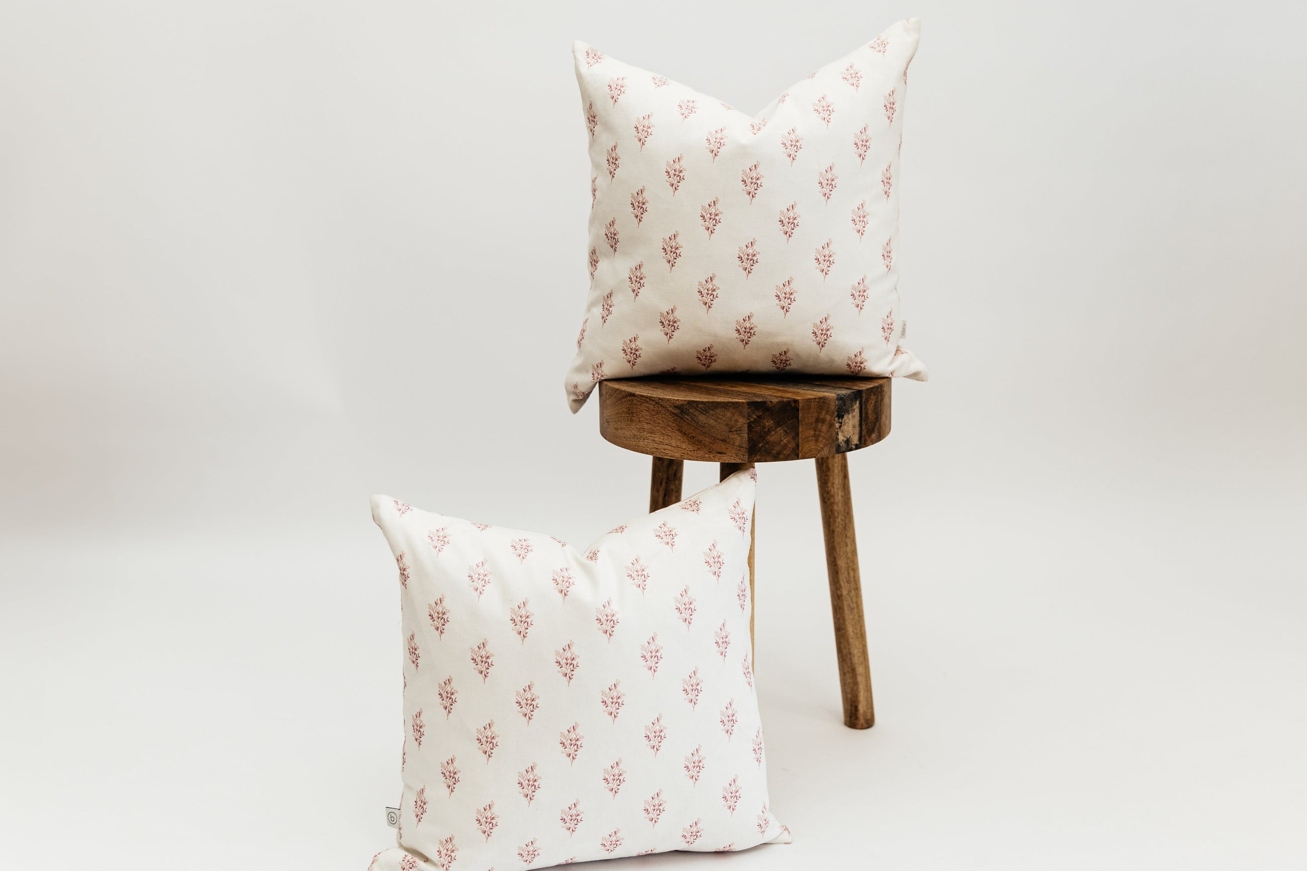 Two Ella pink floral pillows, one on a wooden stool and one leaning against the stool, both positioned in front of a white backdrop