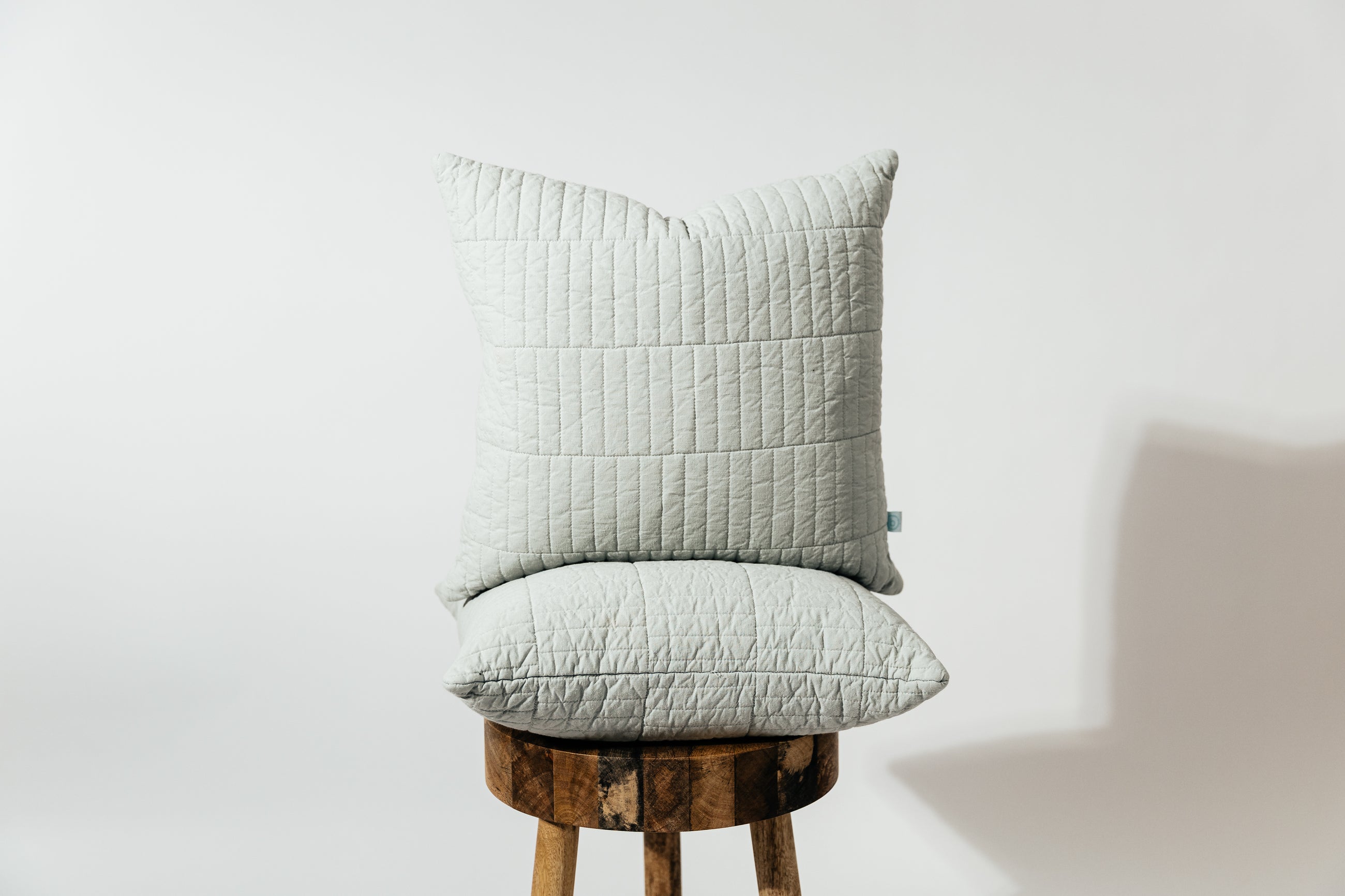 Two blue pillows stacked on top of one another on a wood stool with a white backdrop.