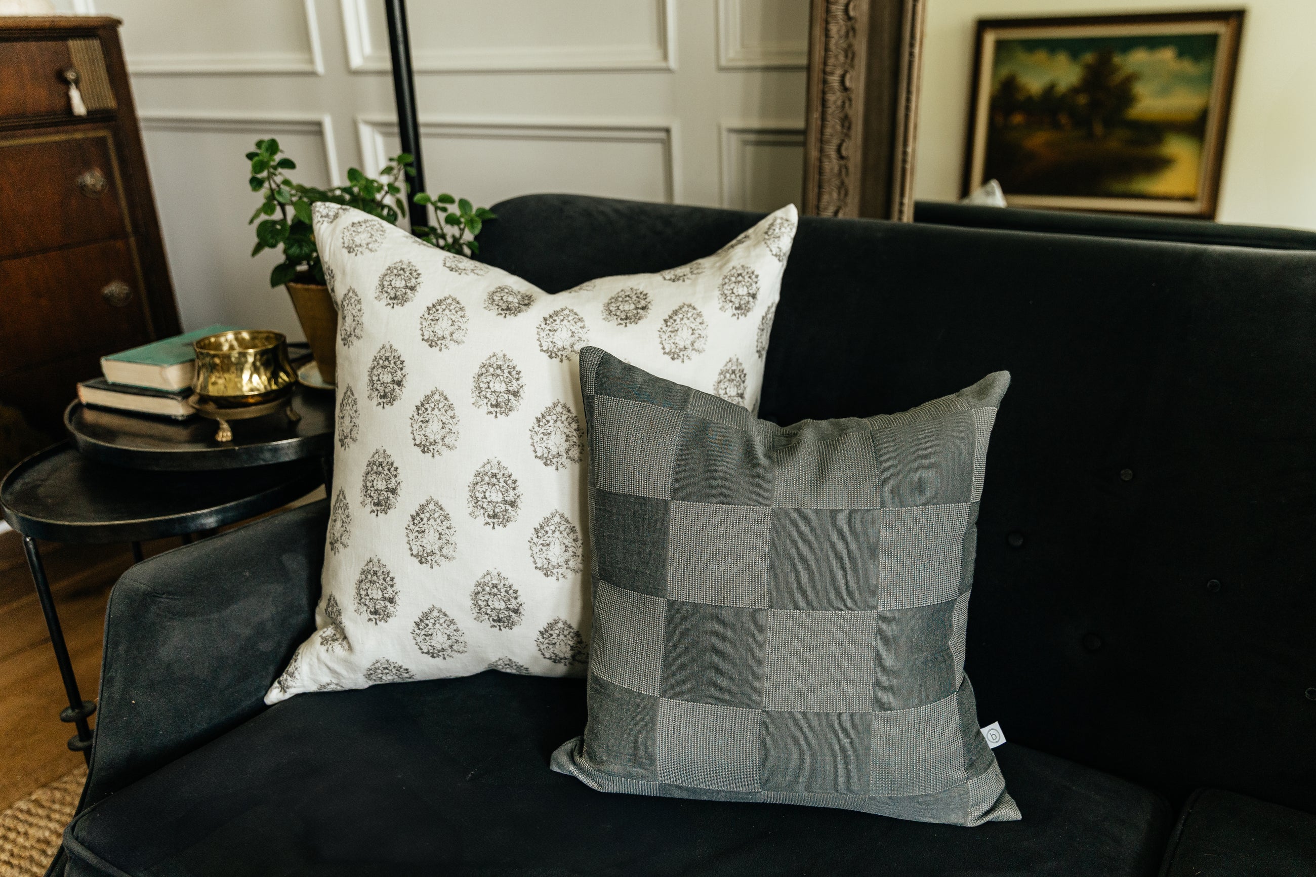 A white and gray floral pillow arranged with a checkerboard gray pillow on a black velvet couch in a living room.