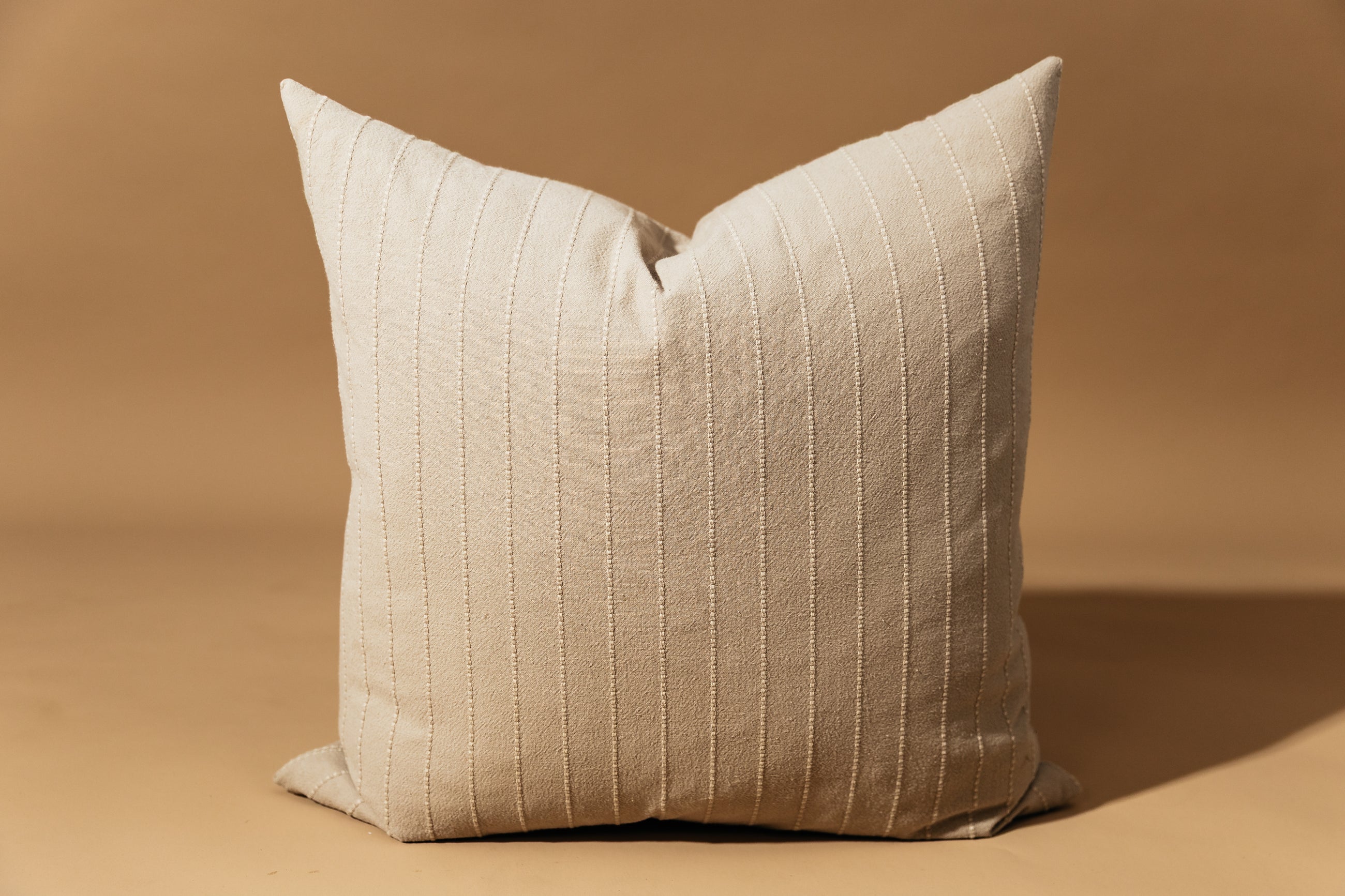 A neutral striped euro sham with vertical stitching arranged in front of a brown backdrop