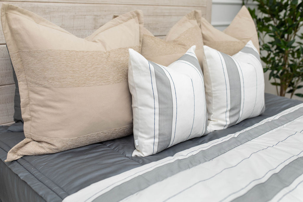 A close up view of a bed made with gray zipper bedding and an assortment of neutral and striped throw pillows and blanket.