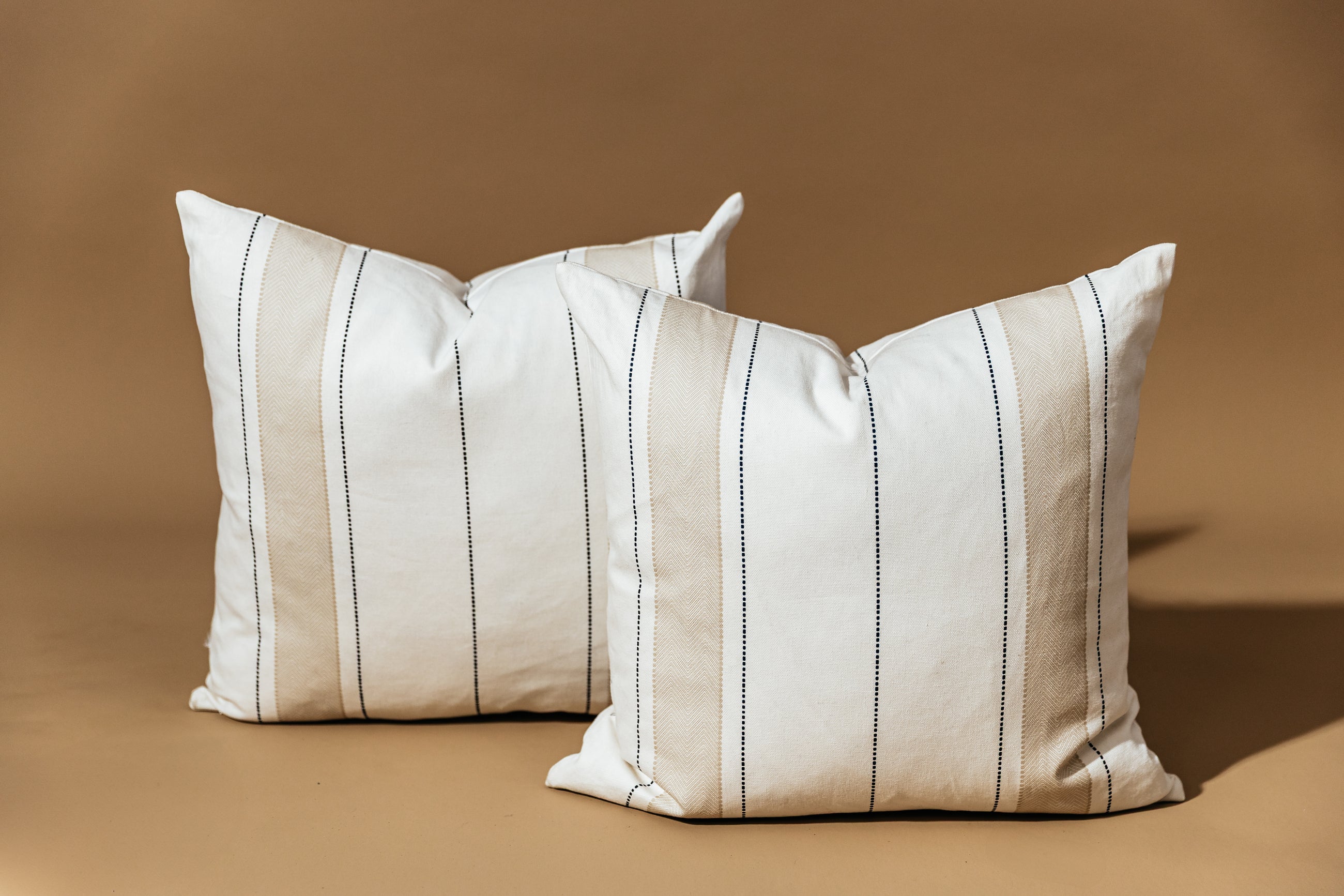 Two neutral striped pillows arranged one in front of the other with a brown backdrop.