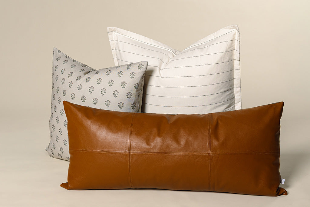A tan, pink, and green blockprint floral pillow, white and black striped pillow, and a leather lumbar on a cream paper backdrop.