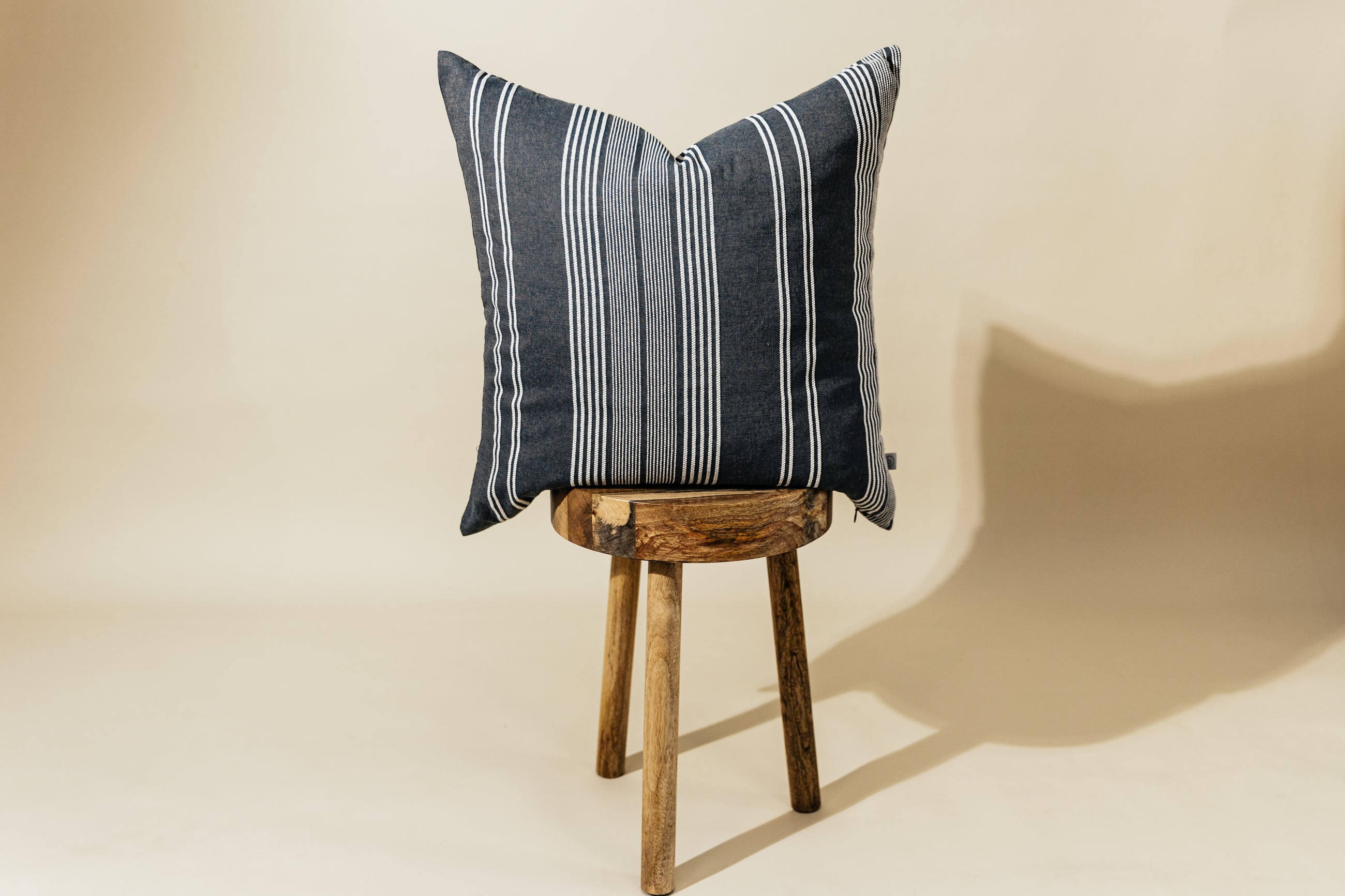 A woven dark gray and white striped euro pillow arranged on a wood stool with a cream background.