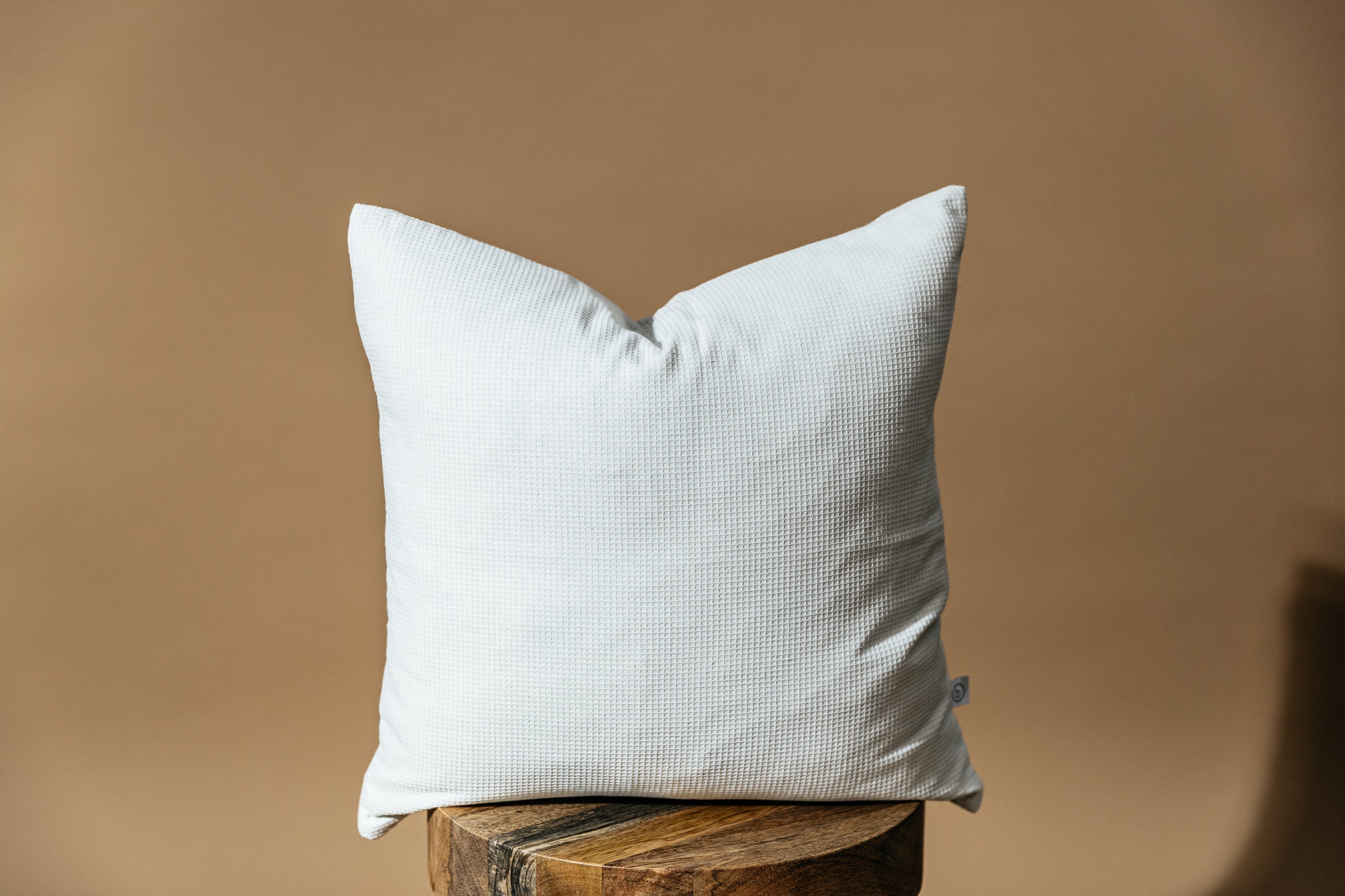 A light gray pillow sitting on a wood stool in front of a brown backdrop.