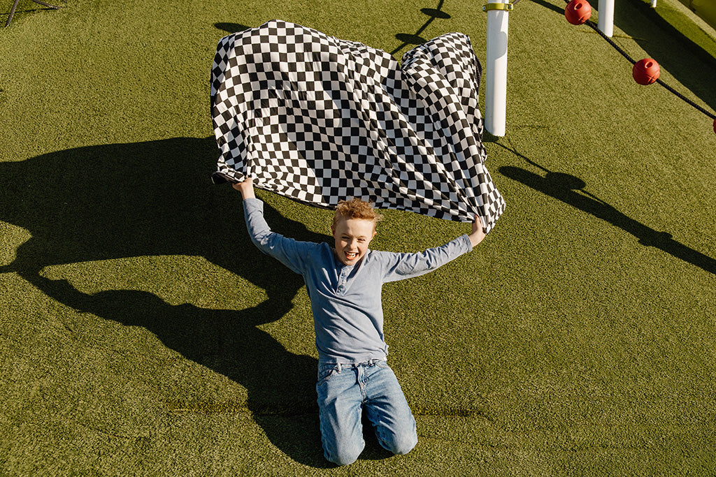 A young boy sliding down a grass hill holding a black and white check blanket. 