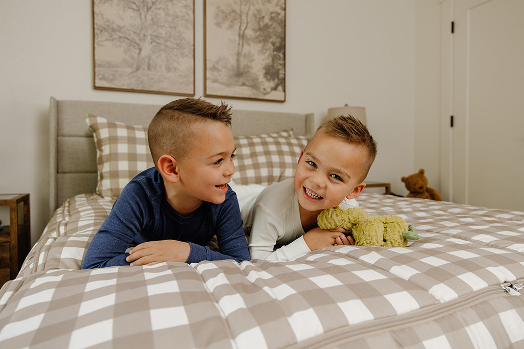 Two young boys laying on their stomachs and smiling on tan gingham bedding.