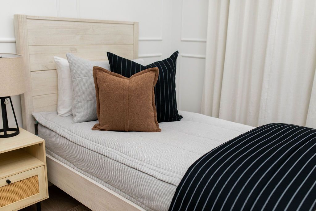 A twin bed made with light gray waffle textured zipper bedding and an assortment of neutral black and brown pillows and a striped black blanket.