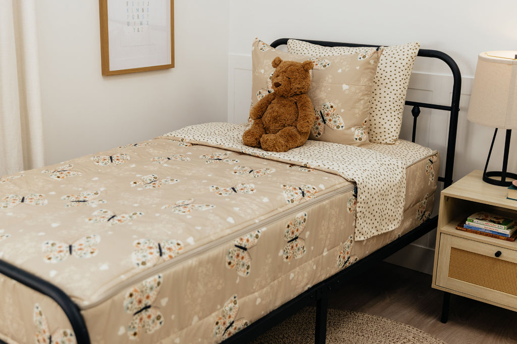 A twin bed made with butterfly zipper bedding, a nightstand beside the bed and a teddy bear propped up against the pillow.
