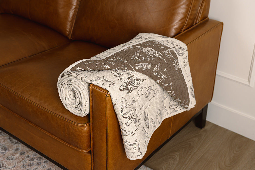 A cream and brown quilted holiday themed blanket on an orange leather couch.
