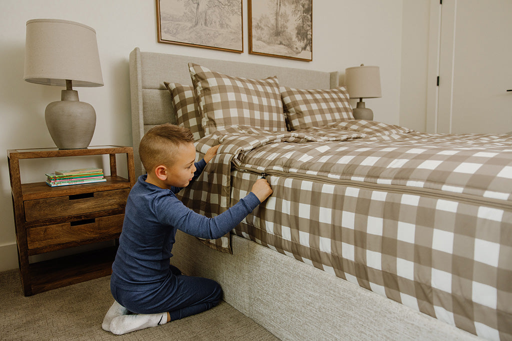 A little boy sitting next to his light brown gingham zipper bedding, zipping his bed up. 