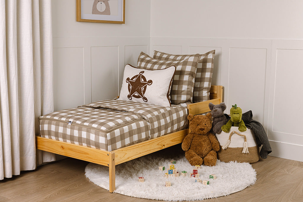 A wooden toddler bed made with tan gingham printed zipper bedding and a stuffed animal leaning against the pillows. 