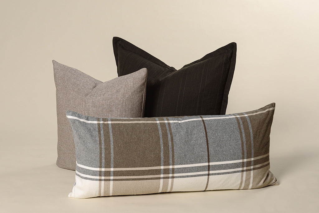 A black knit pillow, brown woven pillow, and plaid lumbar on a cream background.