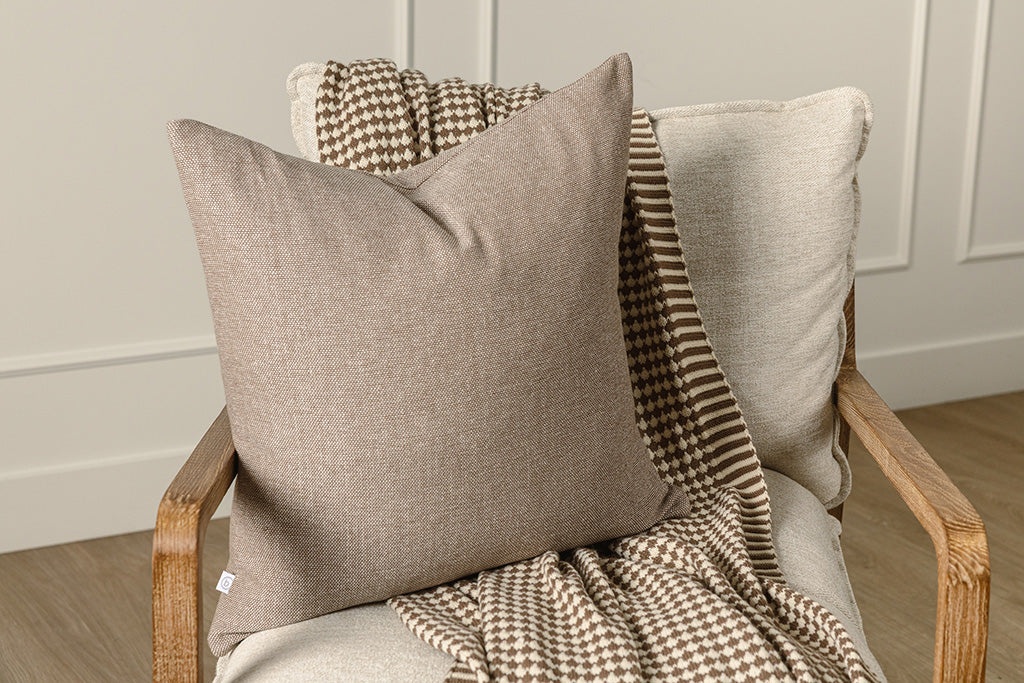 A brown woven pillow and a brown and white scallop knit throw on a wooden and oat colored chair. 