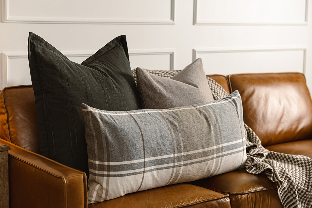 A black knitted pillow, brown woven pillow, plaid lumbar, and a brown and white scallop knit throw on a leather couch.