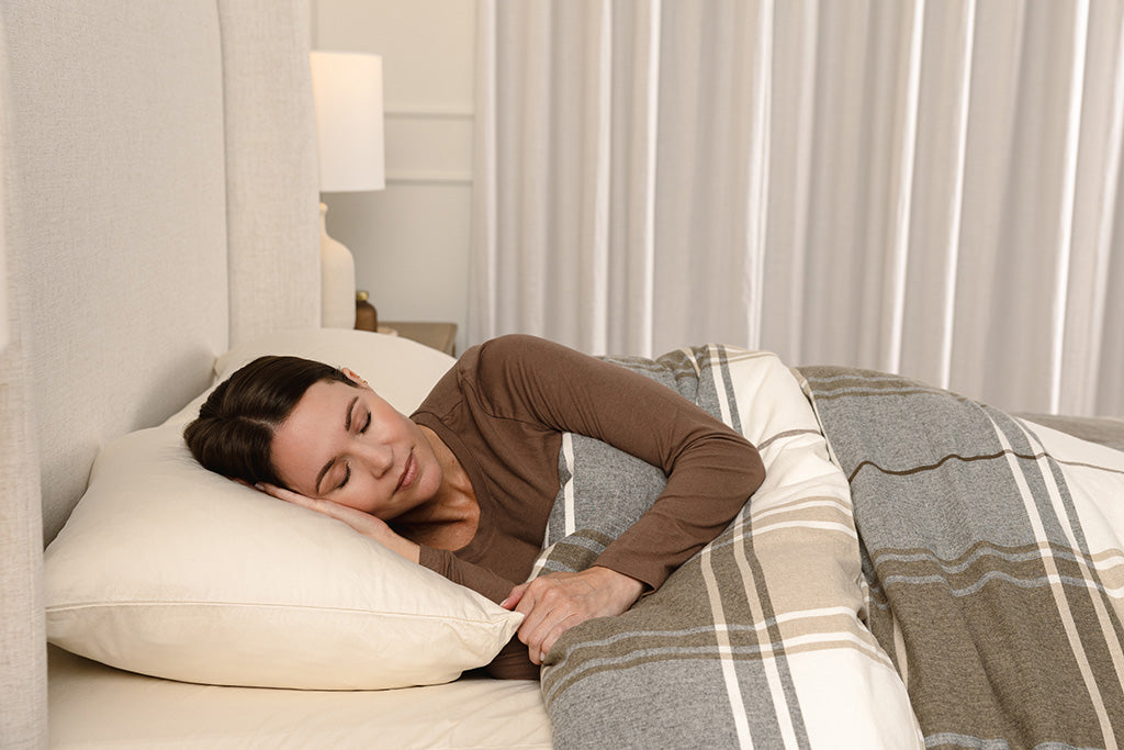 A woman sleeping in a bed with ivory bed sheets and a brown, gray, and white plaid duvet.