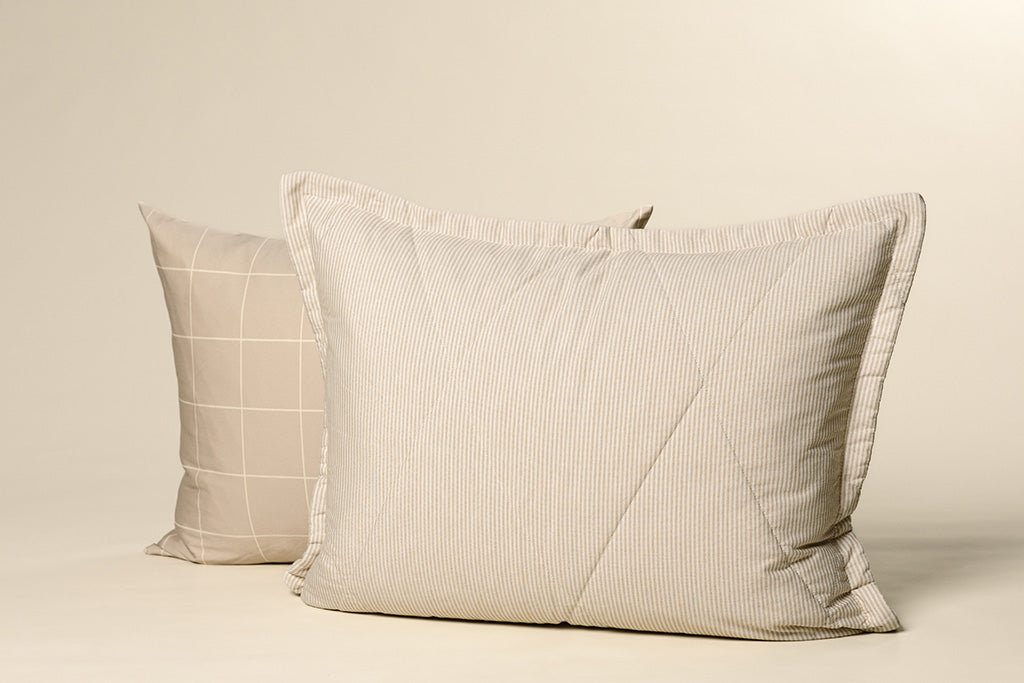 A tan and white grid pillowcase with a white and tan striped quilted sham on a cream backdrop.