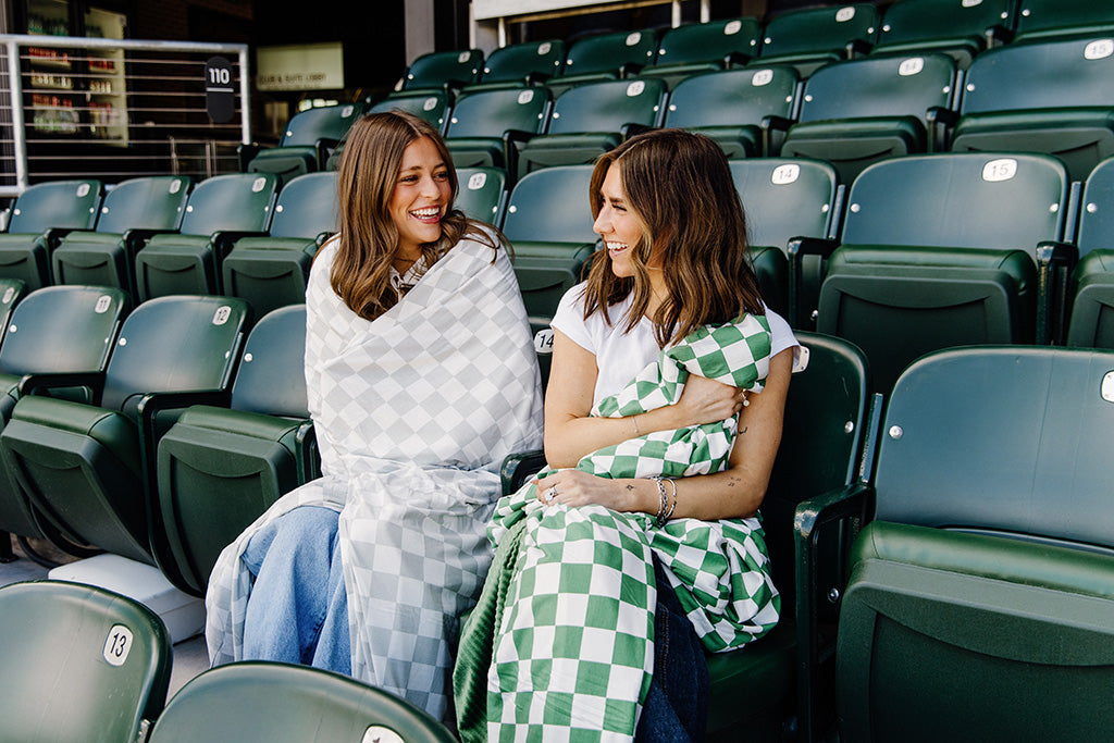 Two women sitting in the stands of a baseball stadium while one is wrapped in a grey and white check blanket and the other in a green and white check blanket.