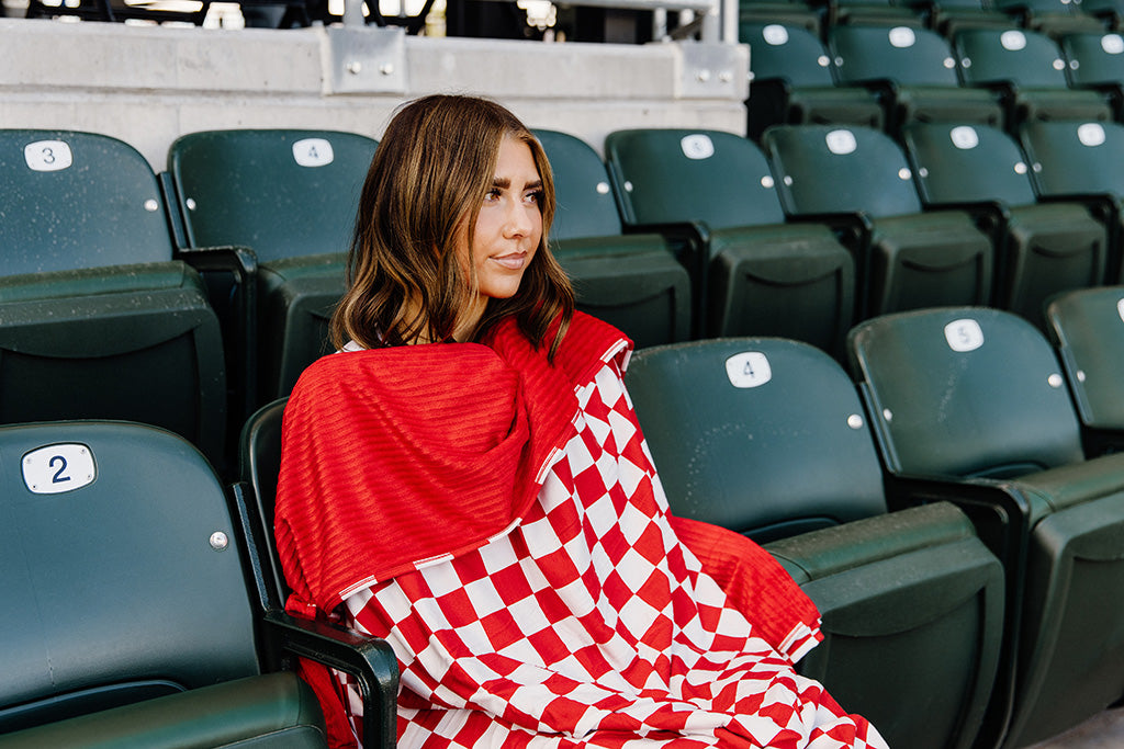 A woman sitting, wrapped in a red and white check blanket in a baseball stadium. 