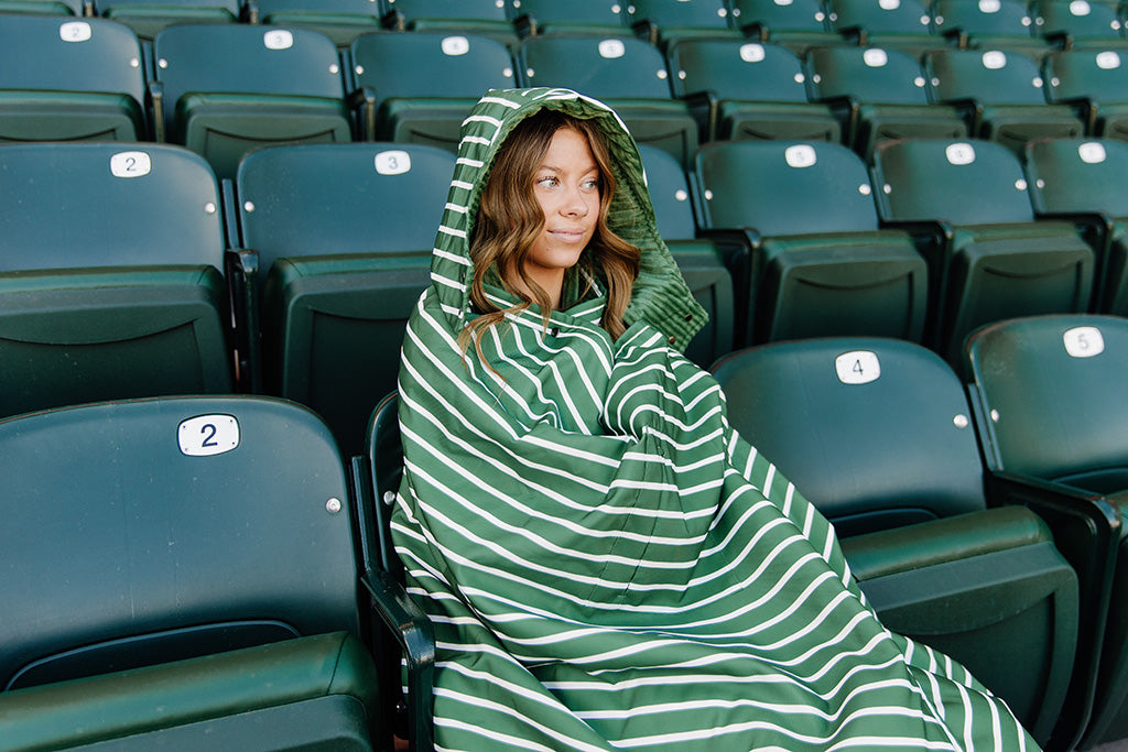 A woman sitting in a baseball stadium wrapped in a green and white striped blanket.