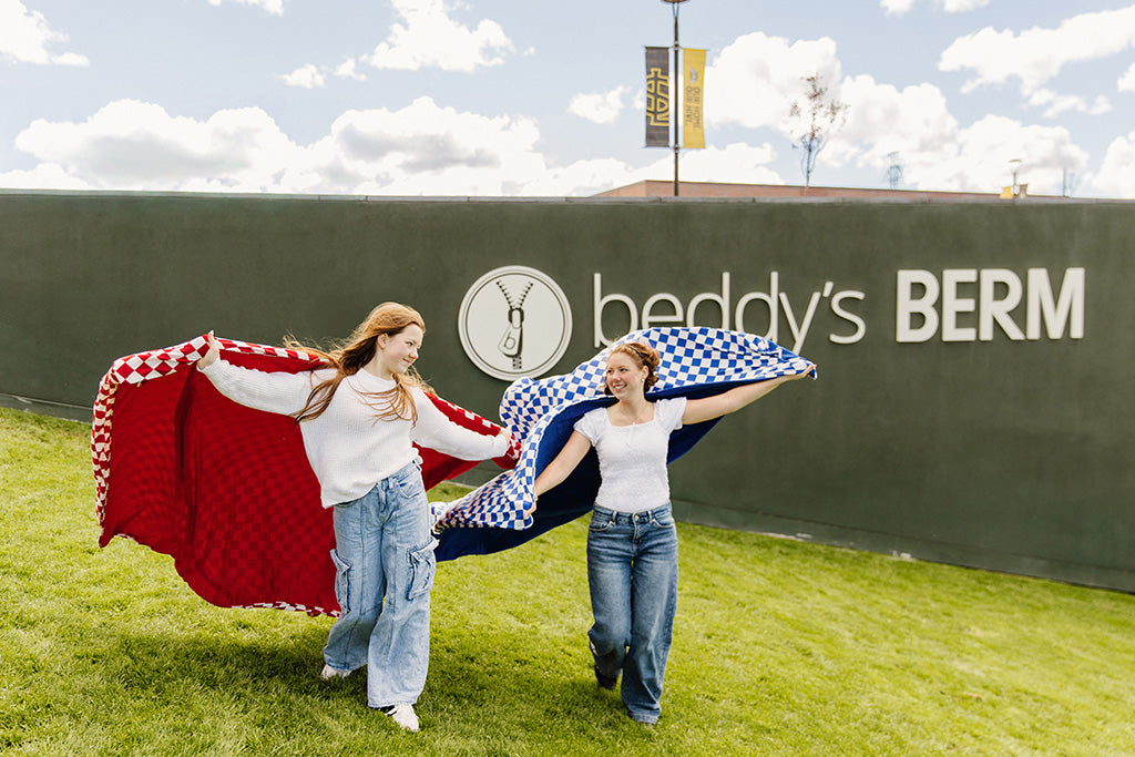 Two girls wrapped in different colored check blankets while running in a grass field. 