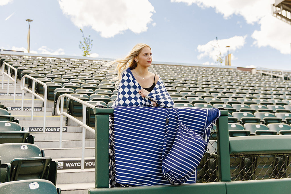 A woman standing in a baseball stadium wrapped in a navy and white check blanket.
