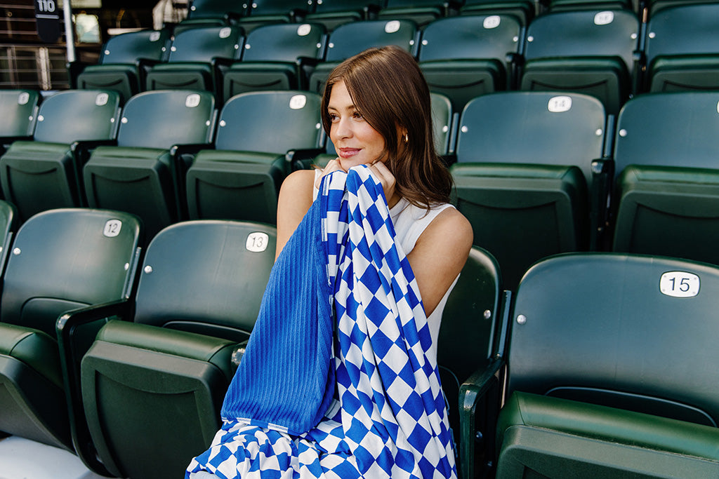 A woman sitting in a baseball stadium while holding her blue and white check blanket close.  
