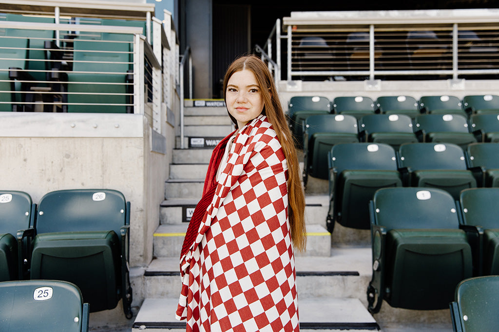 A woman standing, wrapped in a maroon and white check blanket in a baseball stadium. 