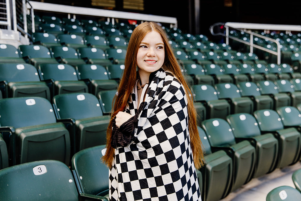 A woman standing, wrapped in a black and white check blanket in a baseball stadium. 