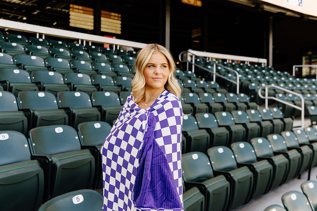A woman standing, wrapped in a purple and white check blanket in a baseball stadium. 