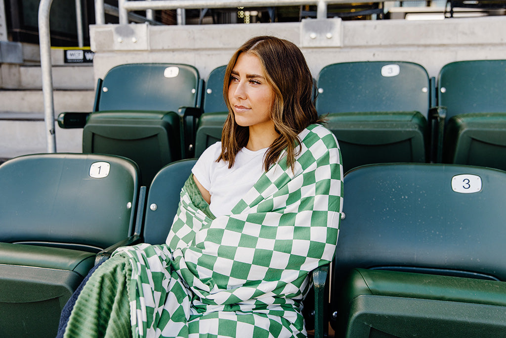 A woman sitting, wrapped in a green and white check blanket in a baseball stadium. 