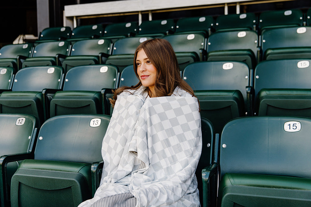 A woman sitting, wrapped in a grey and white check blanket in a baseball stadium. 