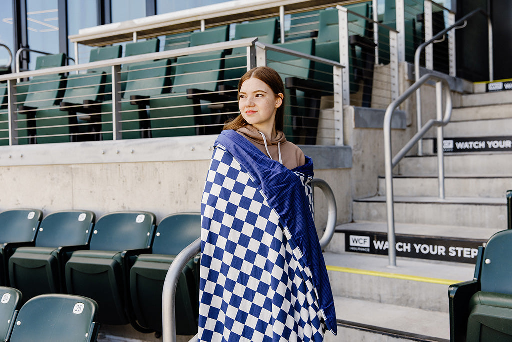 A woman standing, wrapped in a navy and white check blanket in a baseball stadium. 