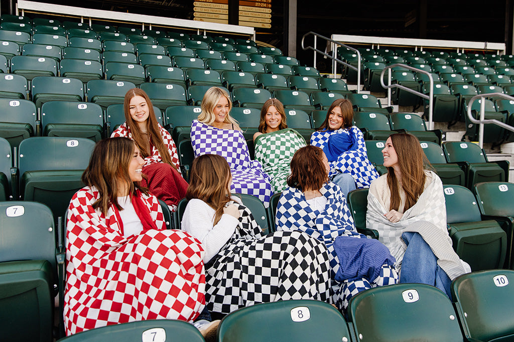 Women sitting wrapped in different colored checked blankets in a baseball stadium, while smiling at each other. 