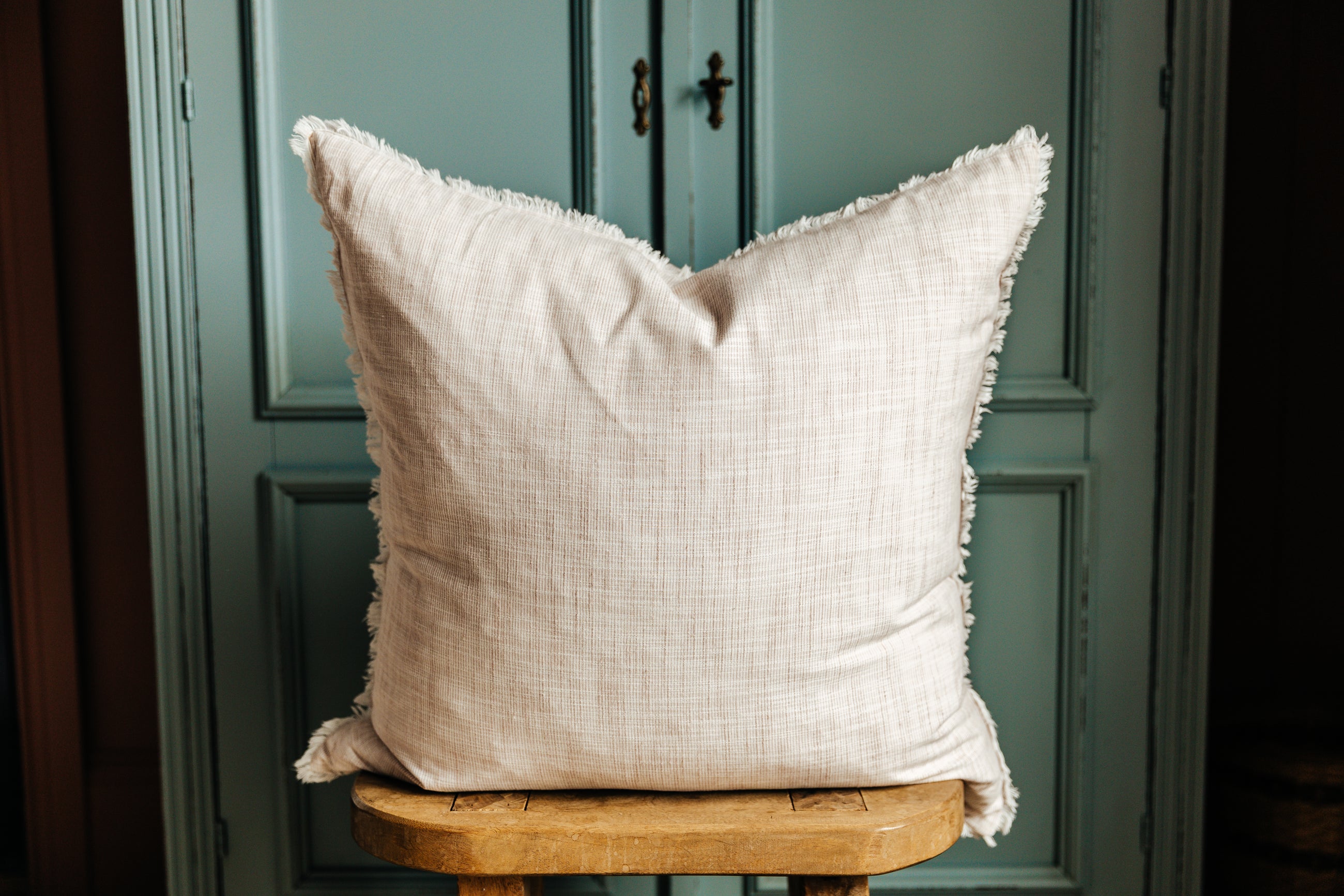 A light pink woven striped euro pillow arranged on a wood stool with blue-green cabinets behind it.