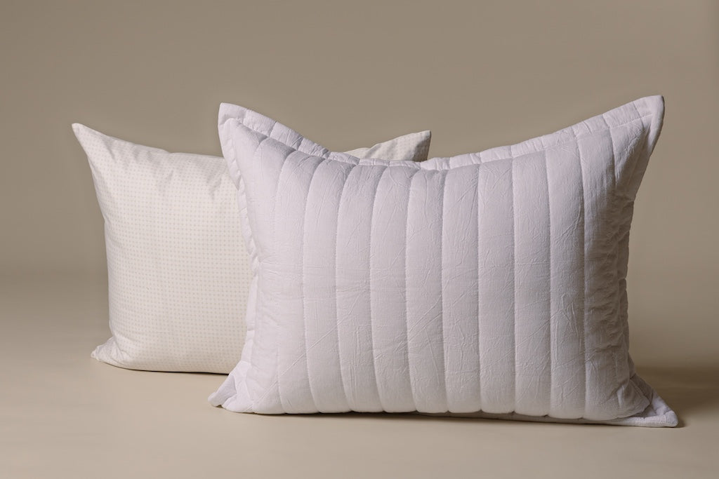 A white and tan crossed pillow and a white striped knitted sham on a cream backdrop.