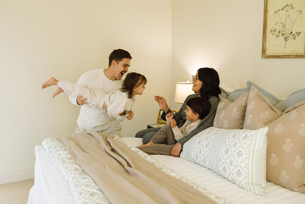 A family snuggling and playing on white stripe bedding with tan and floral accessories. 