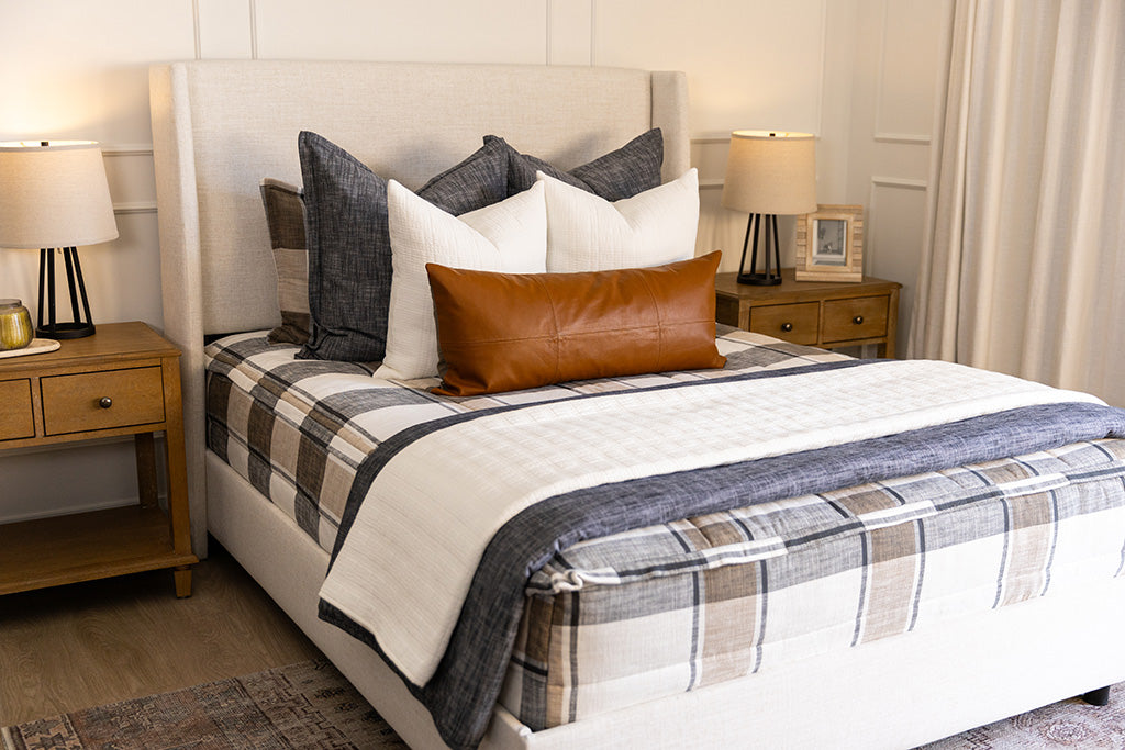 A bed made with charcoal, cream, and tan plaid zipper bedding and an assortment of dark gray and leather throw pillows and matching blankets.