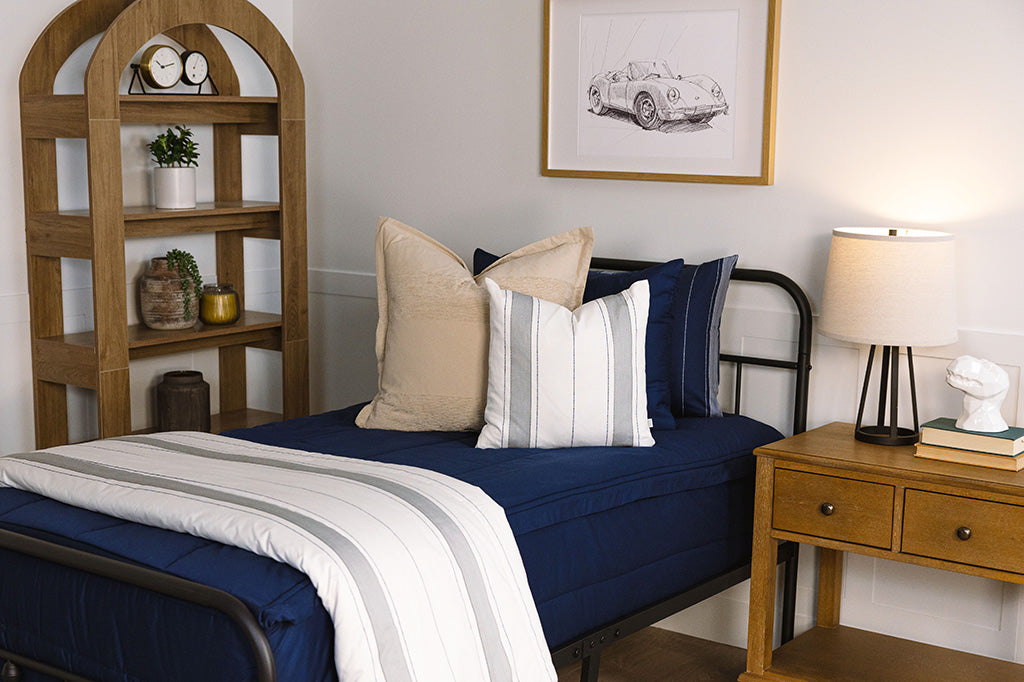 A close up view of a twin bed made with navy blue zipper bedding and neutral and gray striped pillows and blanket.