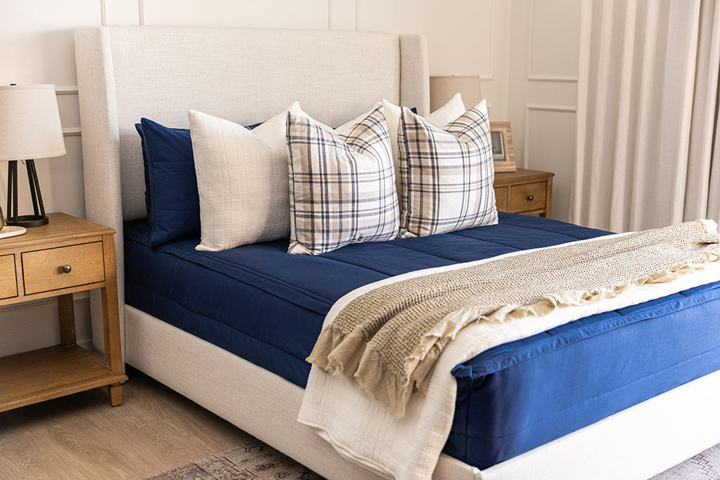 A bed made with navy blue zipper bedding and white and plaid pillows with neutral blankets.