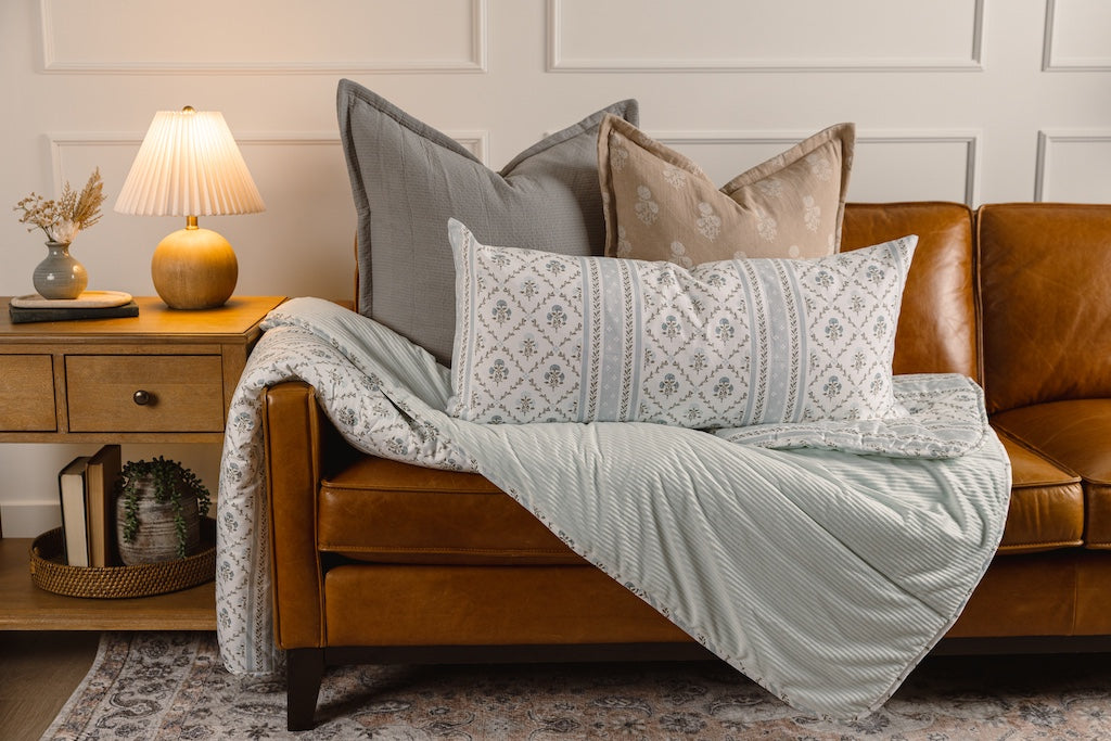 A gray stitched pillow with tan and blue floral pillows, on a blue minky and floral print blanket on an orange leather couch.