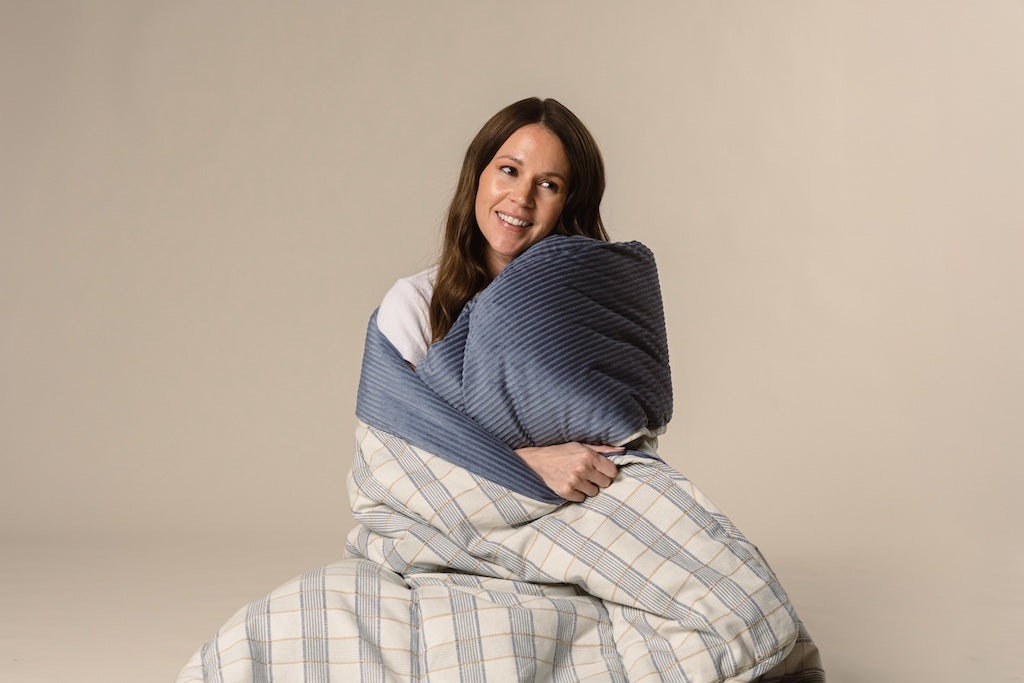 A model wrapped in a blue and yellow plaid stitched blanket with a blue corduroy interior on a cream backdrop.