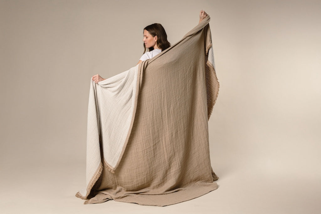 A model holding a light brown and cream with a fringed edge throw on a cream backdrop.