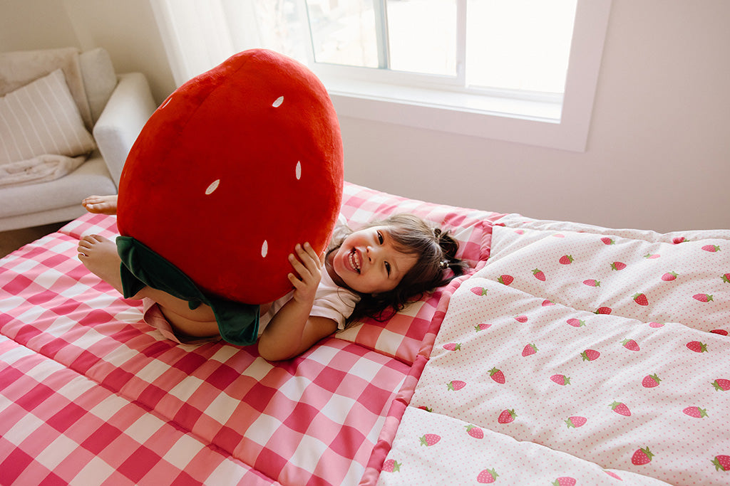 A young girl lying on pink gingham and strawberry print zipper bedding holding a stuffed strawberry pillow.