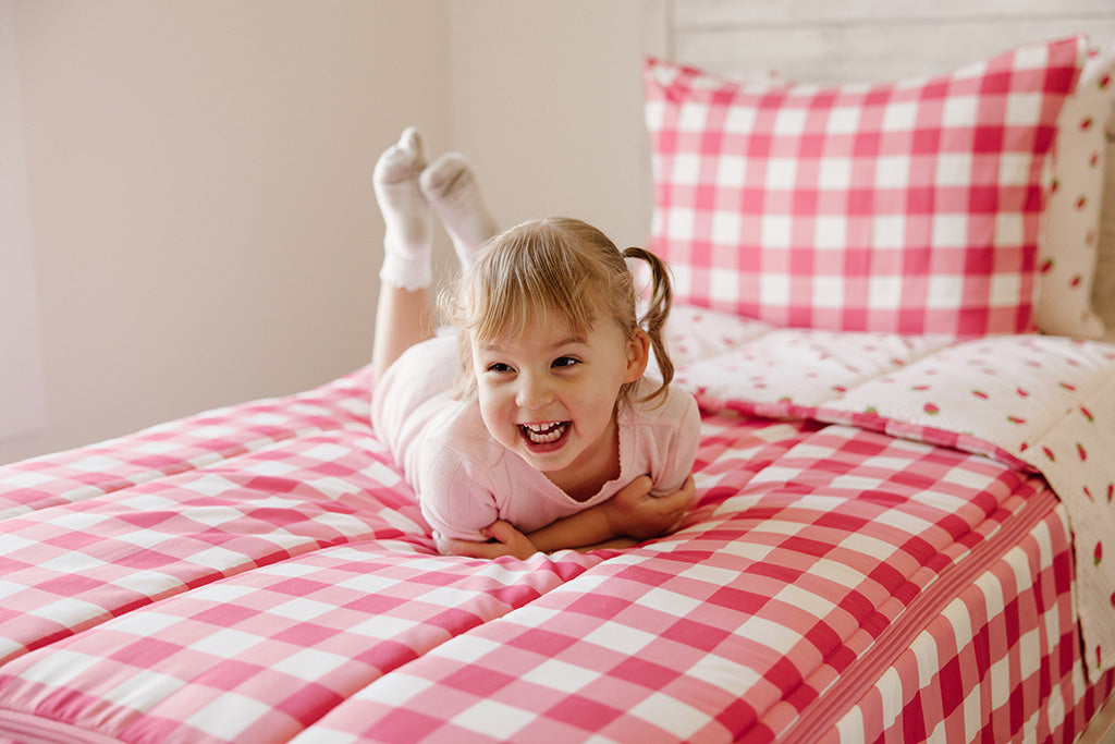 A girl laying down on her stomach and laughing on pink gingham and strawberry print zipper bedding.