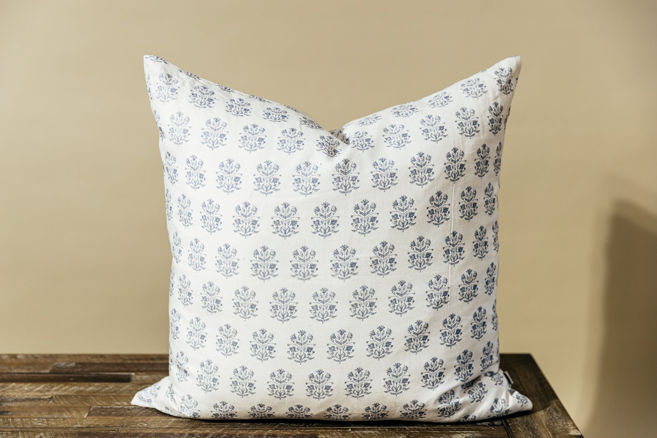 A white euro pillow with a blue floral design arranged on a wood bench with a tan background.