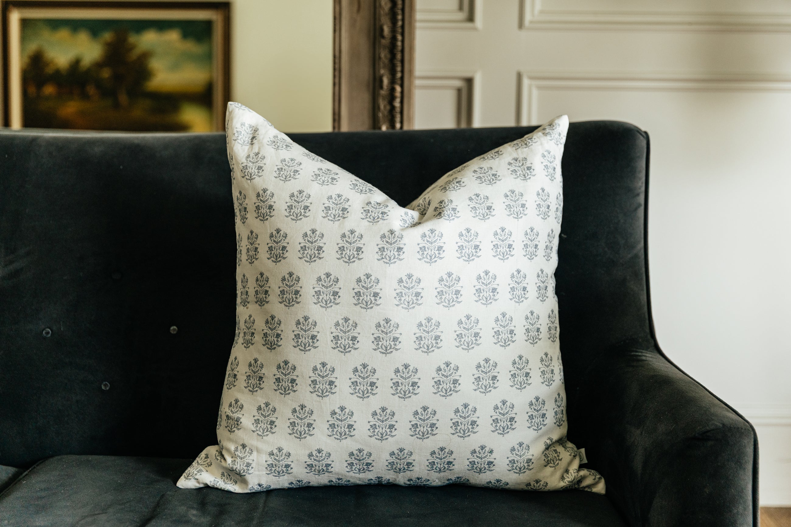 A white euro pillow with a blue floral design arranged on a black velvet couch.