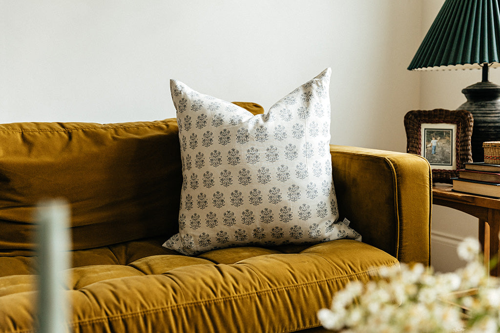 A white euro pillow with a blue floral design arranged on a mustard yellow velvet couch with a side table on the end.